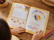 Load image into Gallery viewer, Child's hands holding a childrens journal diary titled 'My Happy Place' on a wooden table with colorful pencils.
